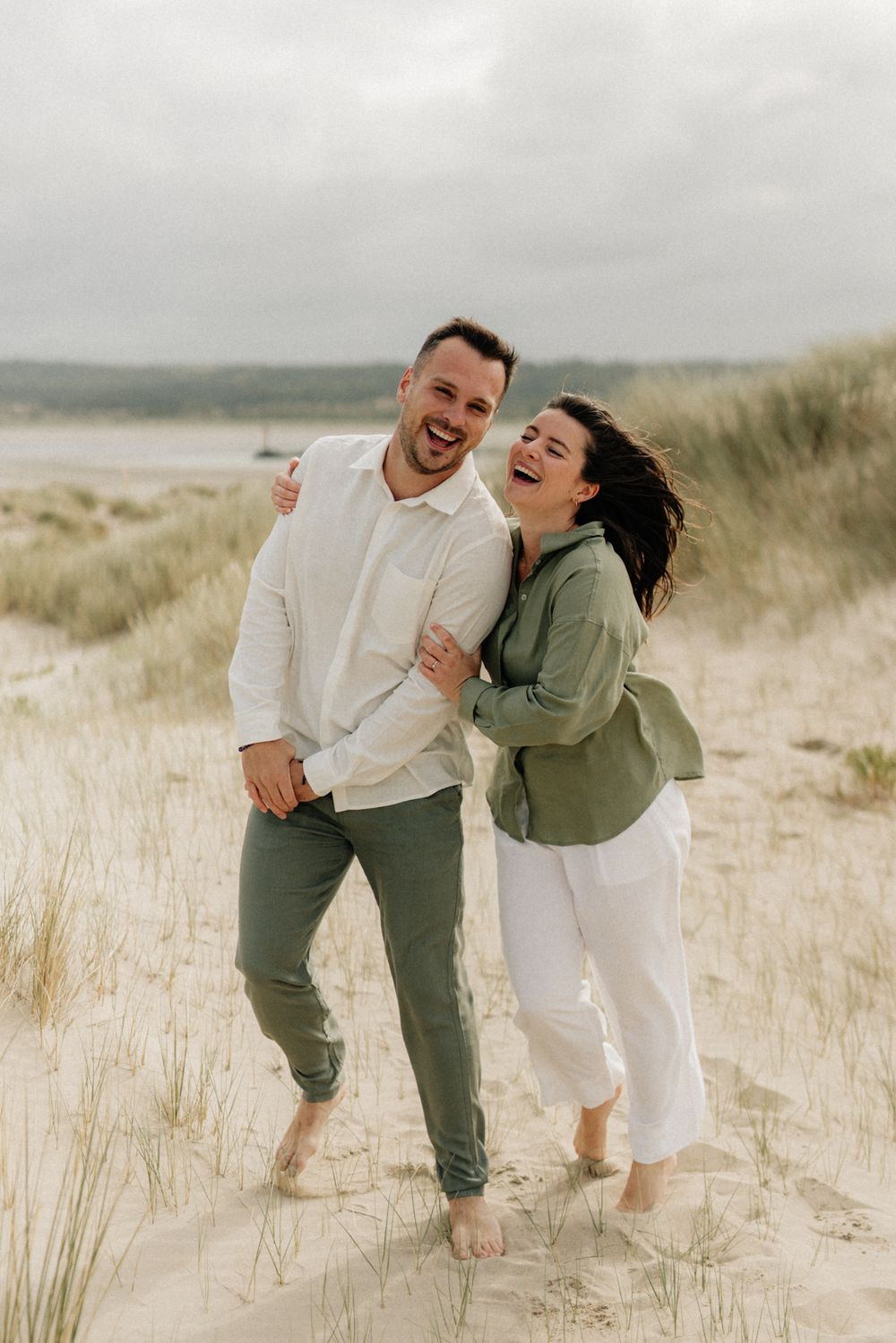 A laughing couple embraces barefoot on sandy beach dunes, man in white shirt and green pants, woman in green jacket and white pants.