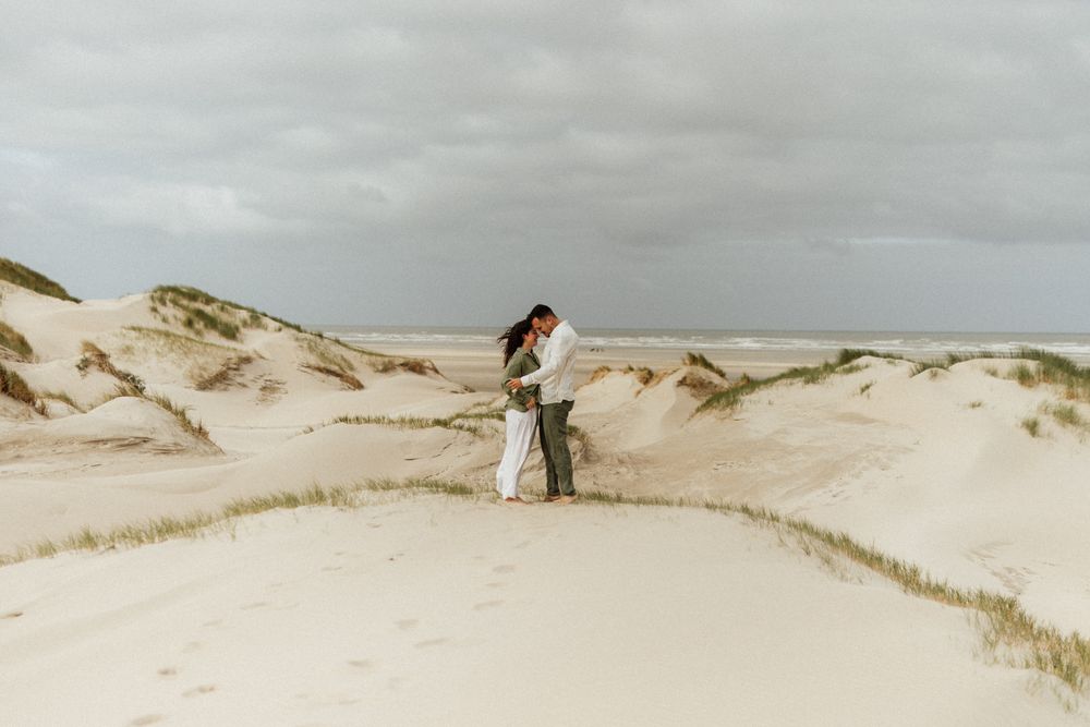 Couple embracing on sandy dunes under cloudy skies, romantic beach wedding photo in natural coastal landscape.