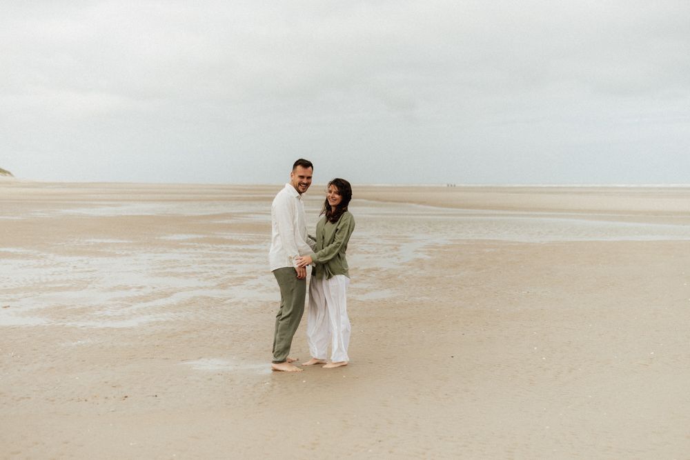 Couple holding hands smiling on a sandy beach with calm ocean waves and overcast sky.