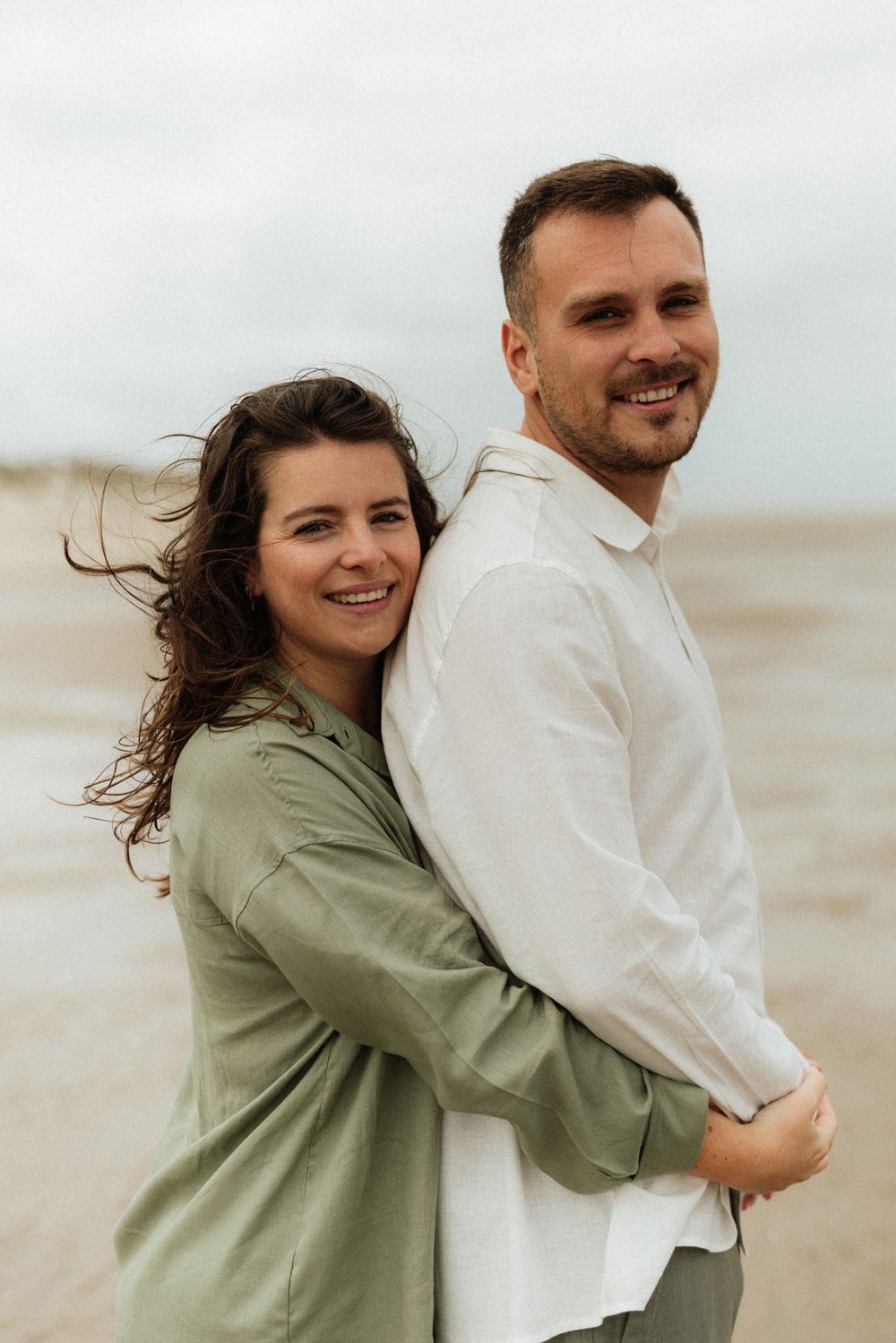 A smiling couple embraces on a windy beach, woman in green shirt hugs man in white linen shirt from behind.