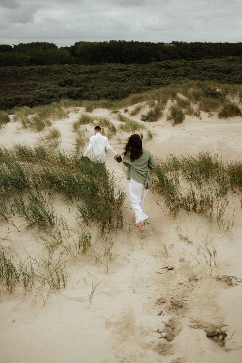 Couple holding hands walking through sandy beach dunes with green grass and trees in the background.