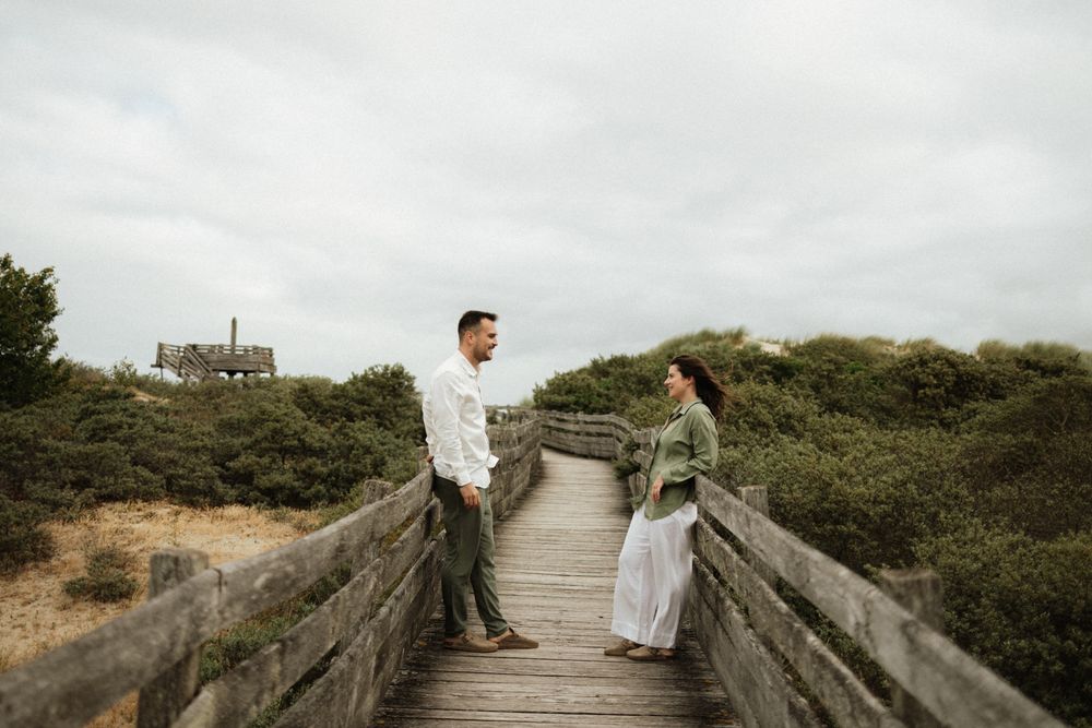 Couple standing face to face on a wooden boardwalk surrounded by green shrubs under an overcast sky.
