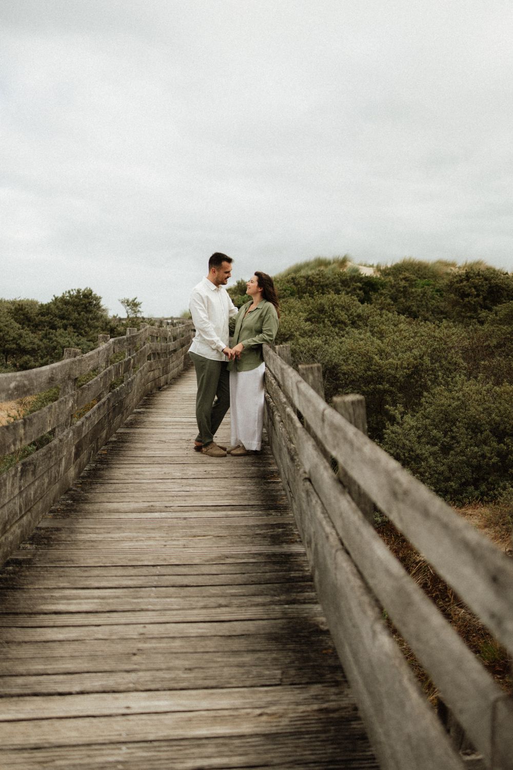 Couple holding hands on a wooden boardwalk surrounded by green shrubs under an overcast sky.
