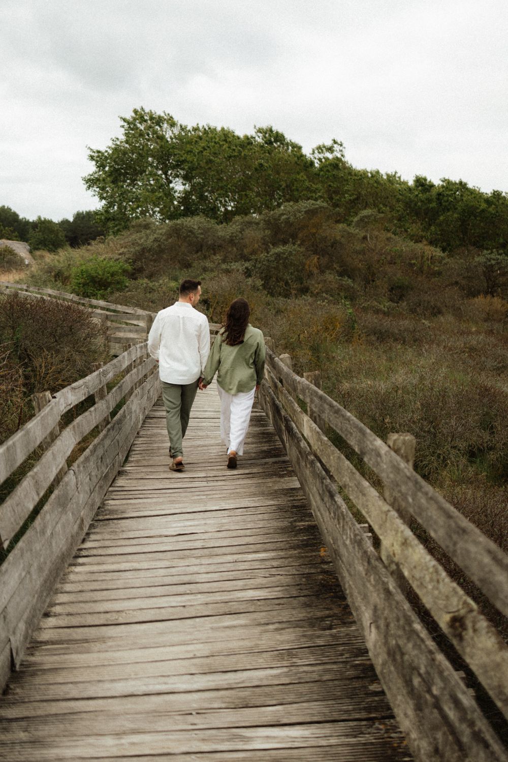 Couple holding hands walking along a wooden boardwalk path through green coastal dunes on a cloudy day.