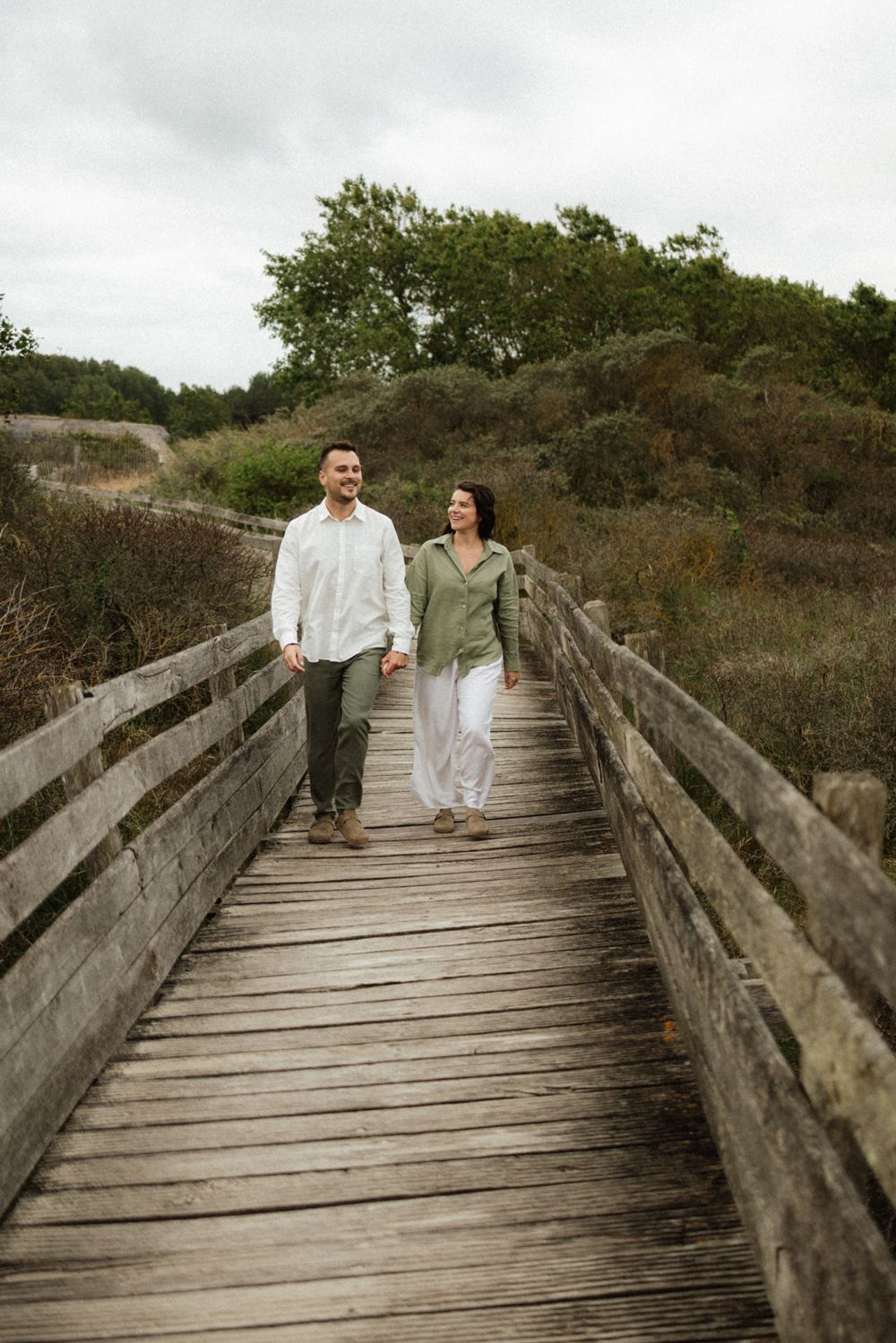 Couple walking hand in hand on a wooden boardwalk path surrounded by green shrubs and trees on a cloudy day.
