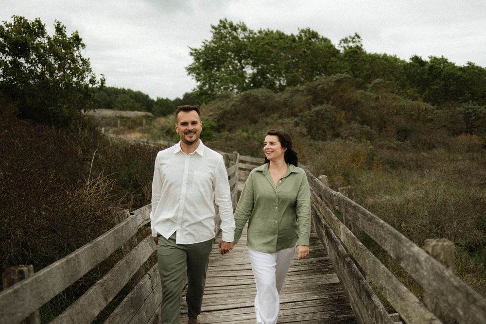 Couple holding hands walking on wooden boardwalk path surrounded by lush greenery on a cloudy day.