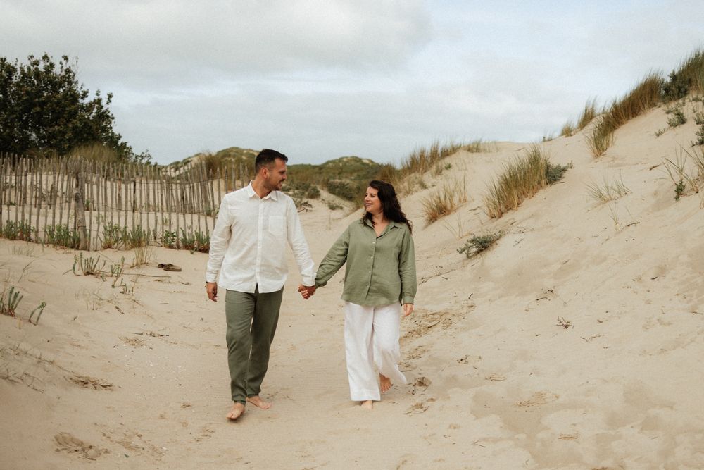 Couple holding hands walking on sandy beach dunes, wearing casual linen outfits, smiling at each other on a cloudy day.