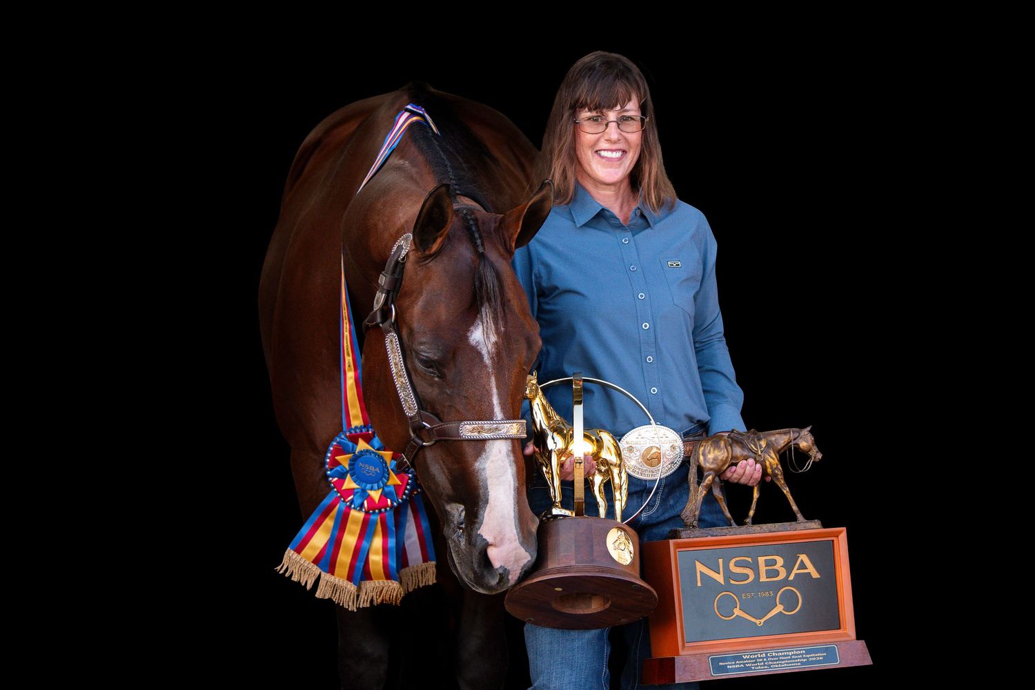 Young woman smiling with bay horse, holding NSBA trophy, ribbons, and awards at equestrian competition.