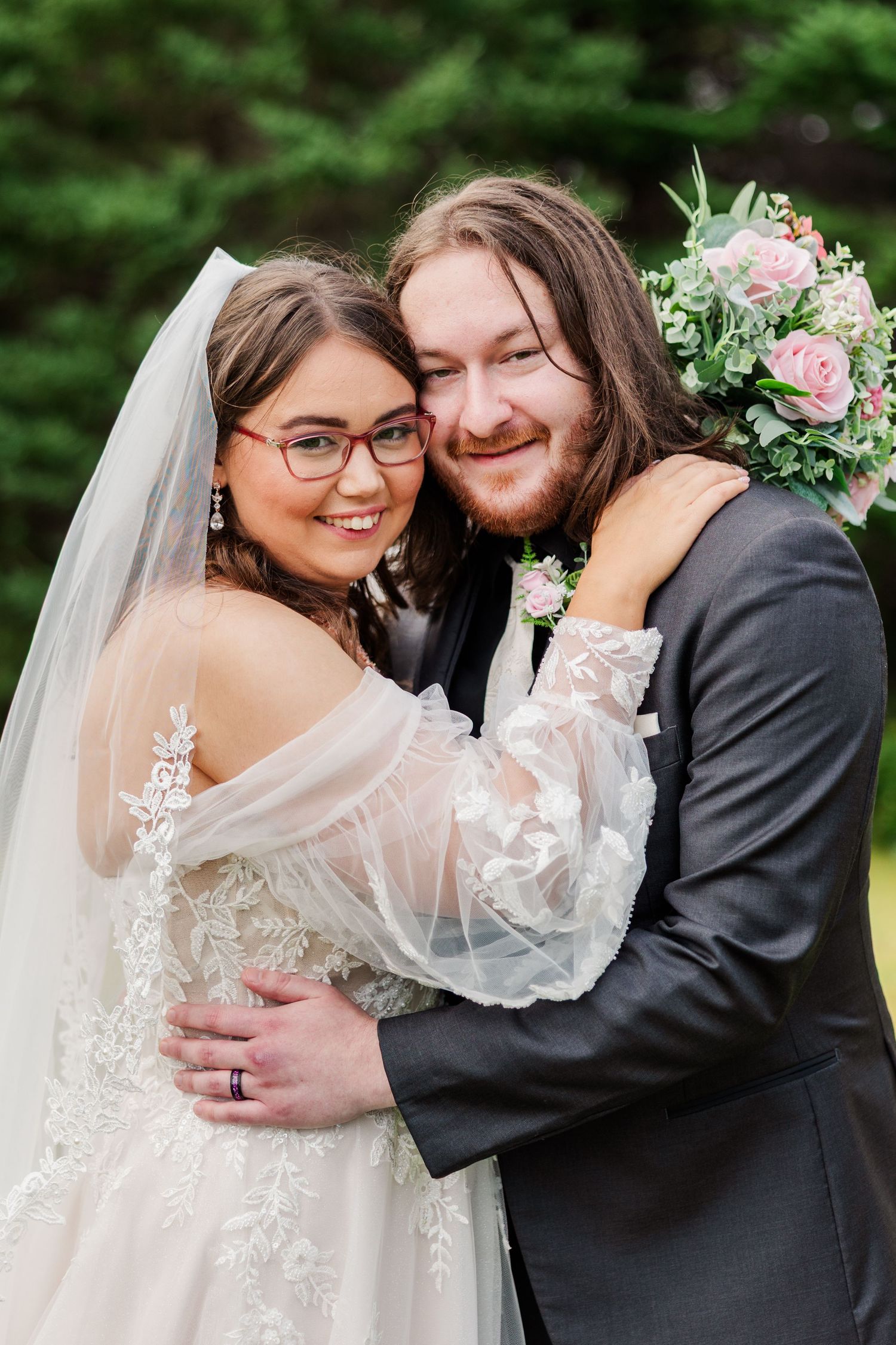 A smiling bride in lace gown and veil embraces her groom in gray suit, posing together outdoors on their wedding day.