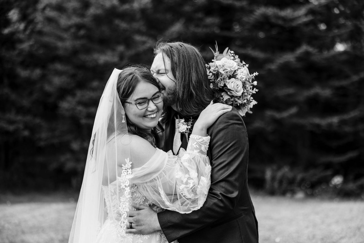 Black and white wedding photo of bride and groom sharing a joyful embrace outdoors, bride holding bouquet.