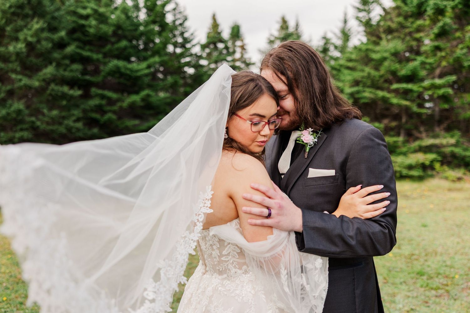 Bride and groom share intimate embrace, her lace veil flowing in wind, surrounded by evergreen trees.
