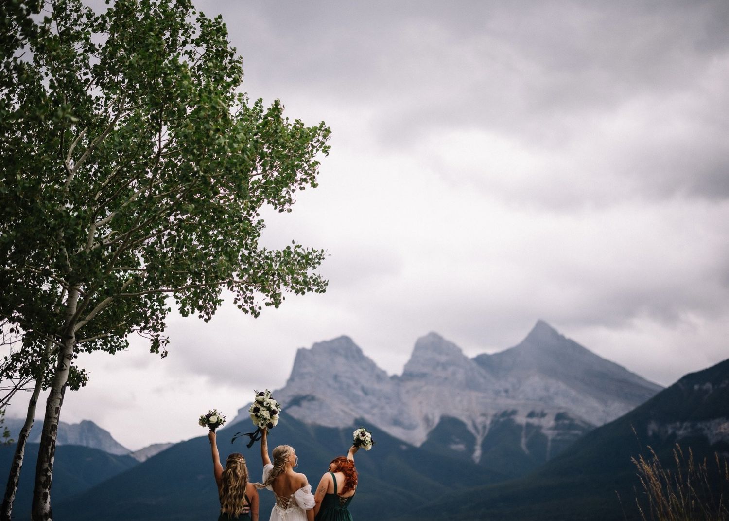 Bride and bridesmaids in green raising their bouquets toward the Three Sisters in Canmore, Alberta