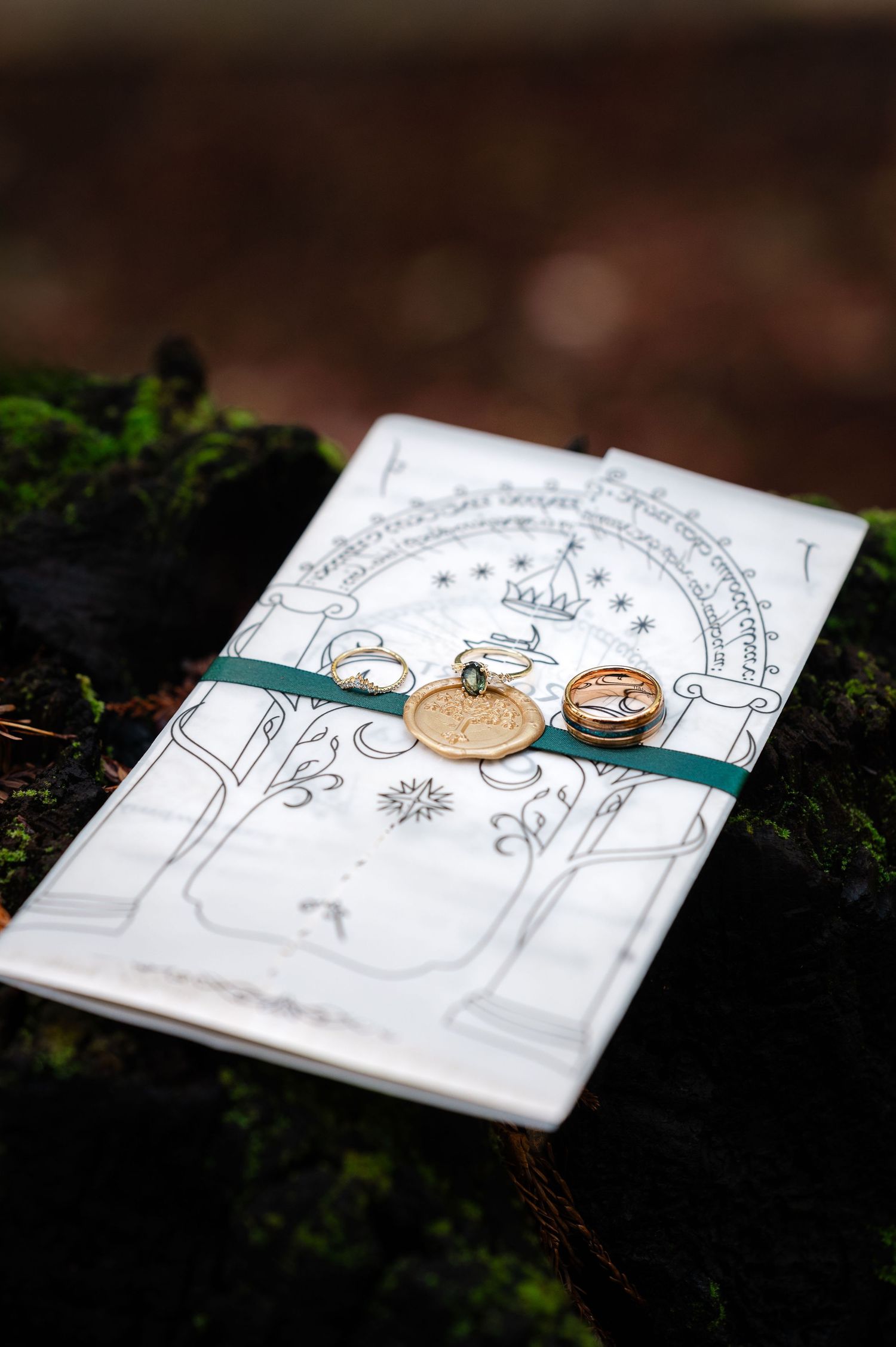 Lord of the Rings themed wedding invitation with gold rings and wax seal on moss-covered surface.