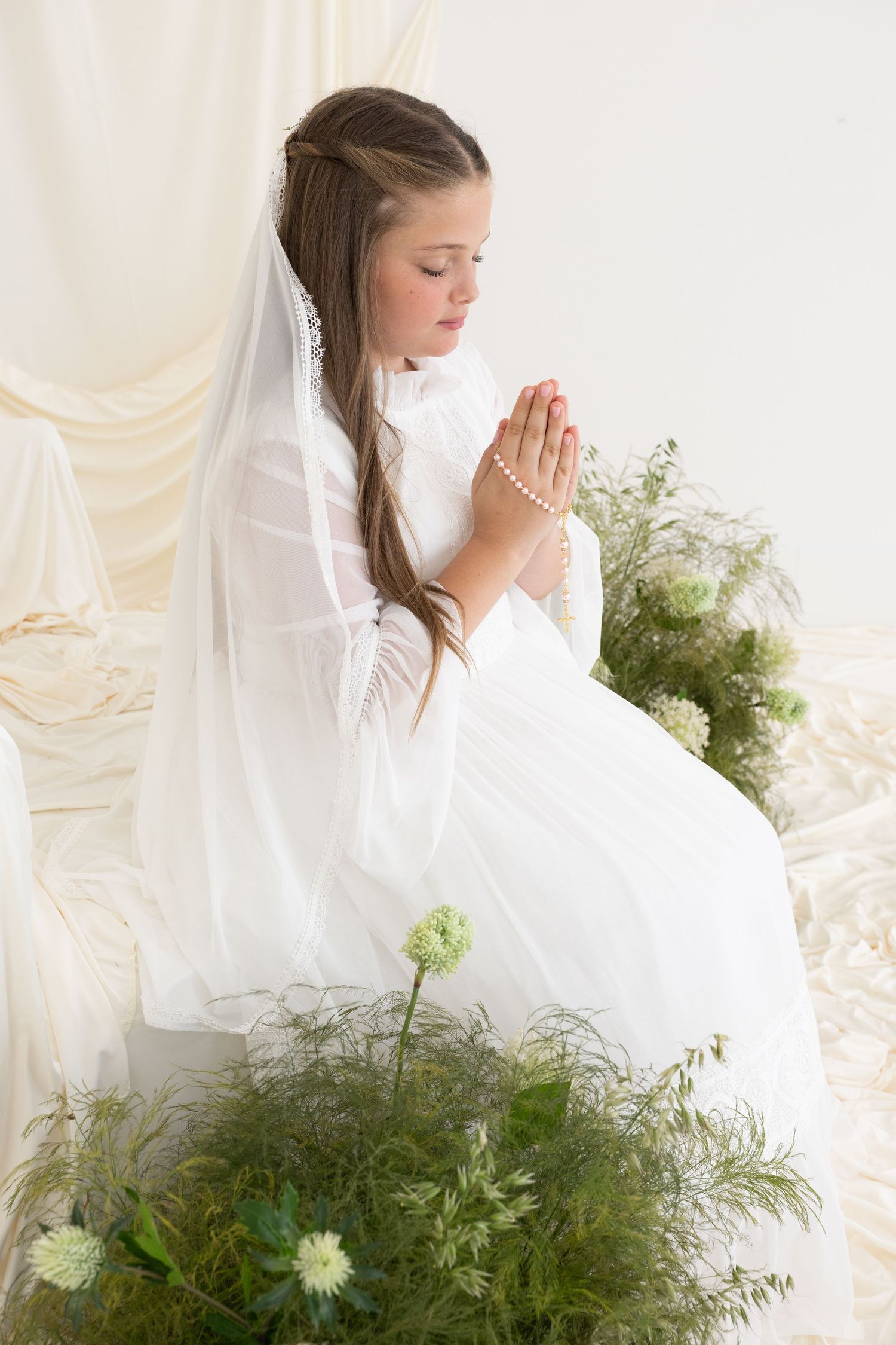 Young girl in white First Communion dress and veil praying with rosary beads, surrounded by green flowers.