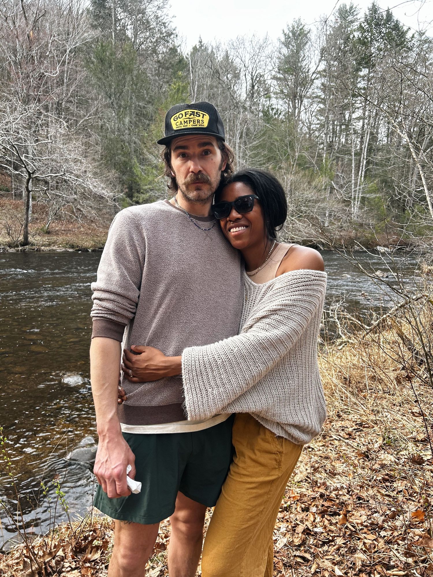 Couple embracing outdoors near a river, surrounded by bare trees and forest in early spring.