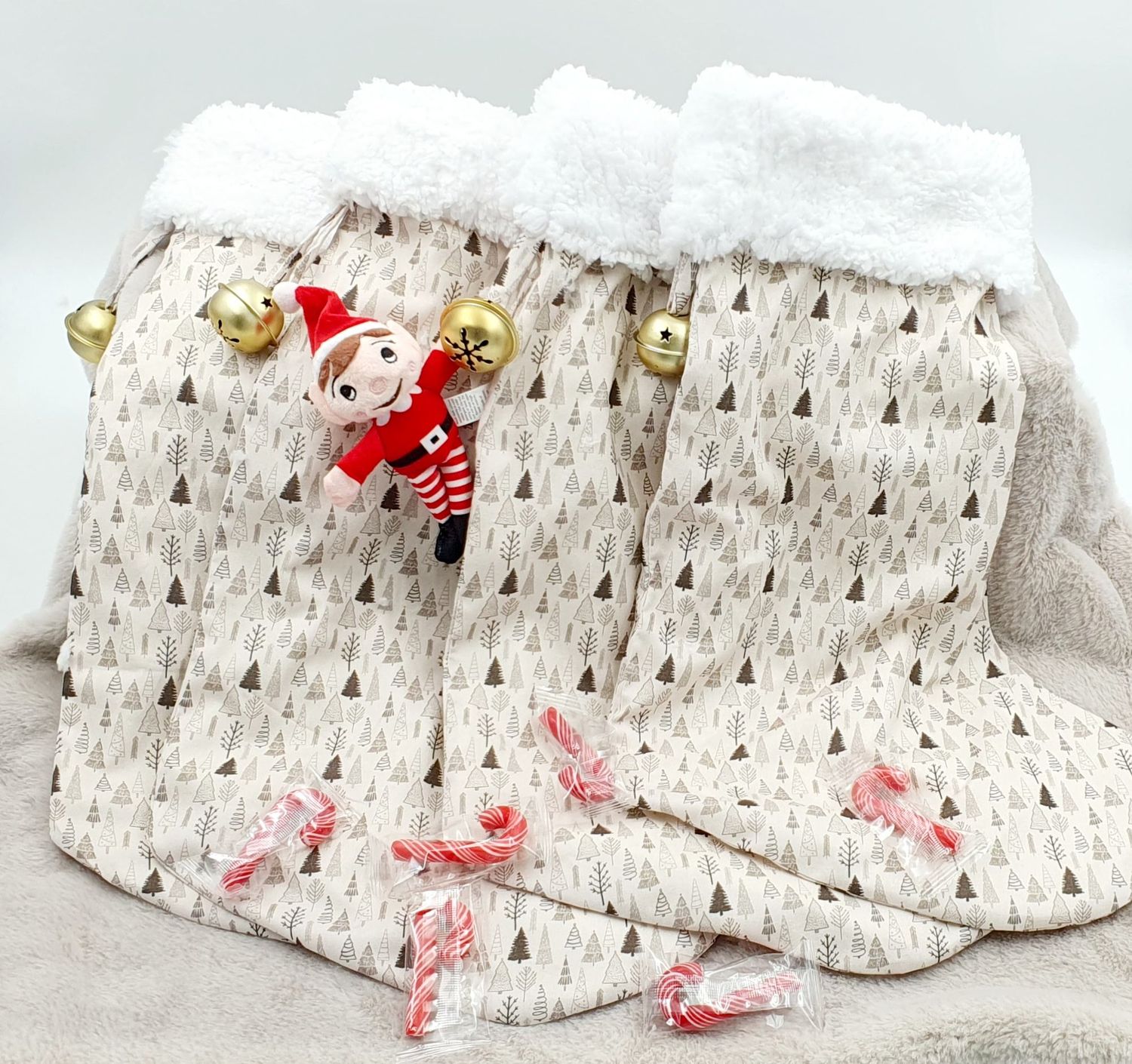 Christmas stockings with white fur trim, gold bells, tree print fabric, an elf doll, and candy canes on a cozy blanket.