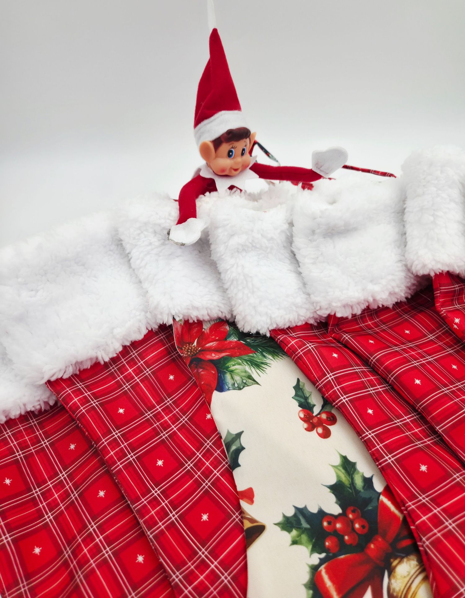 Elf on the Shelf doll resting in red plaid Christmas stockings with white fur trim and holly berry decorations.