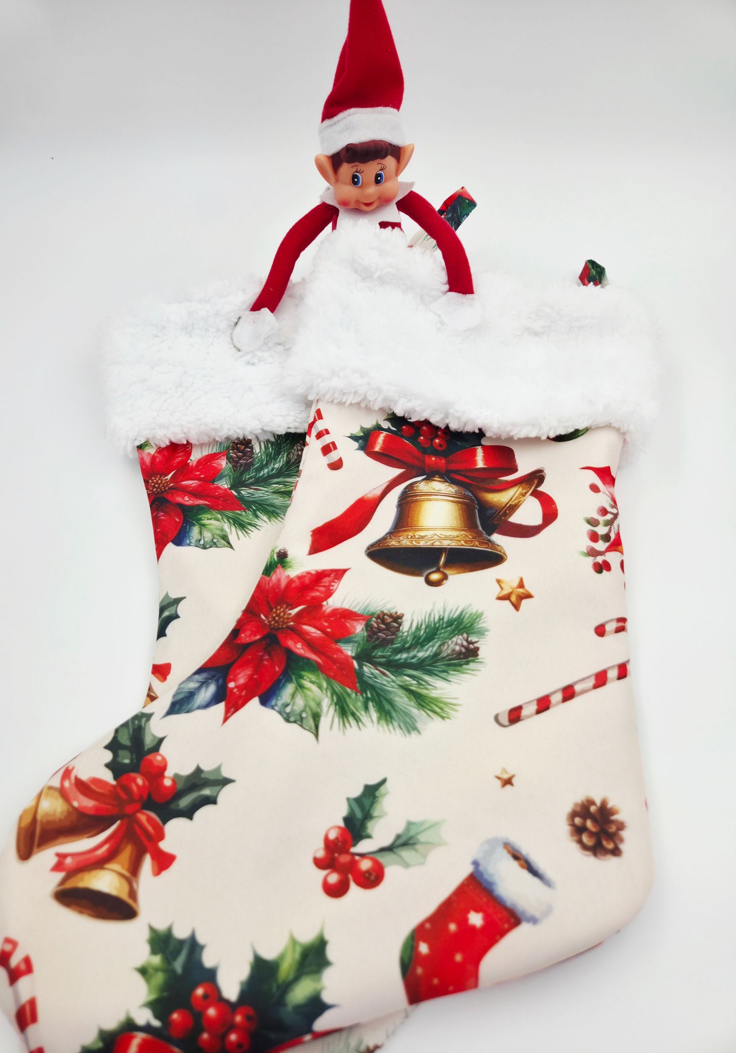 An Elf on the Shelf doll peeking out of a festive Christmas stocking decorated with poinsettias, bells, and holly.