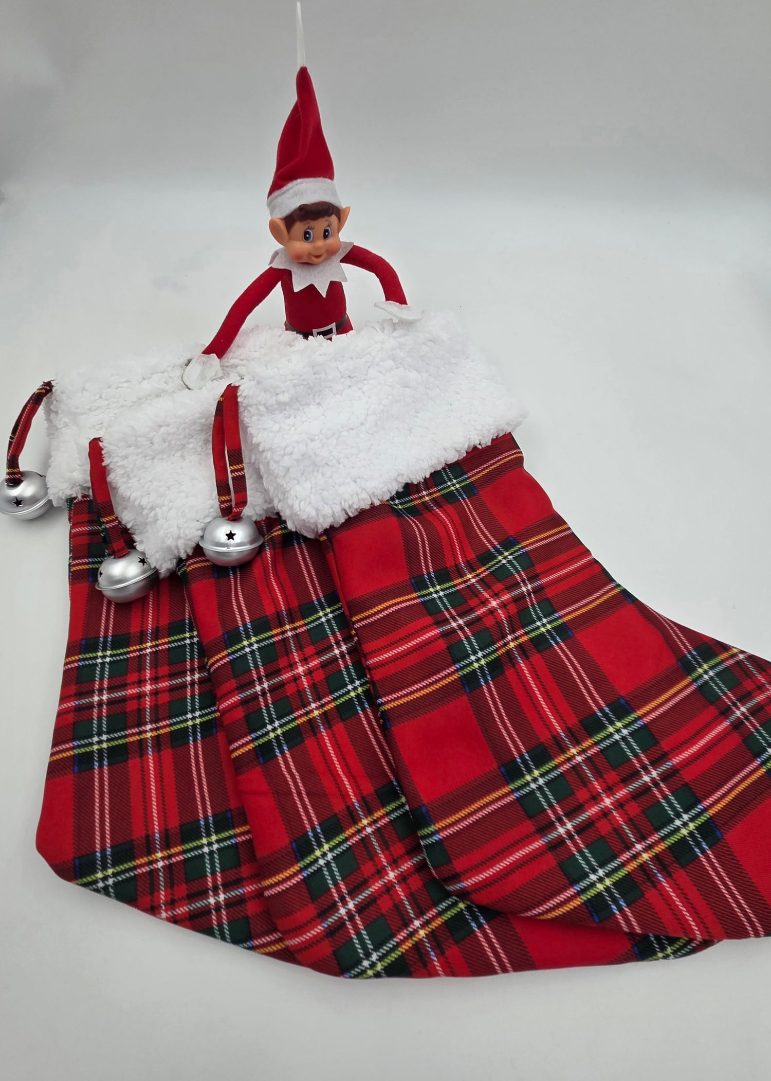 Elf on the Shelf doll sitting in a red plaid Christmas stocking with white fur trim and silver jingle bells.
