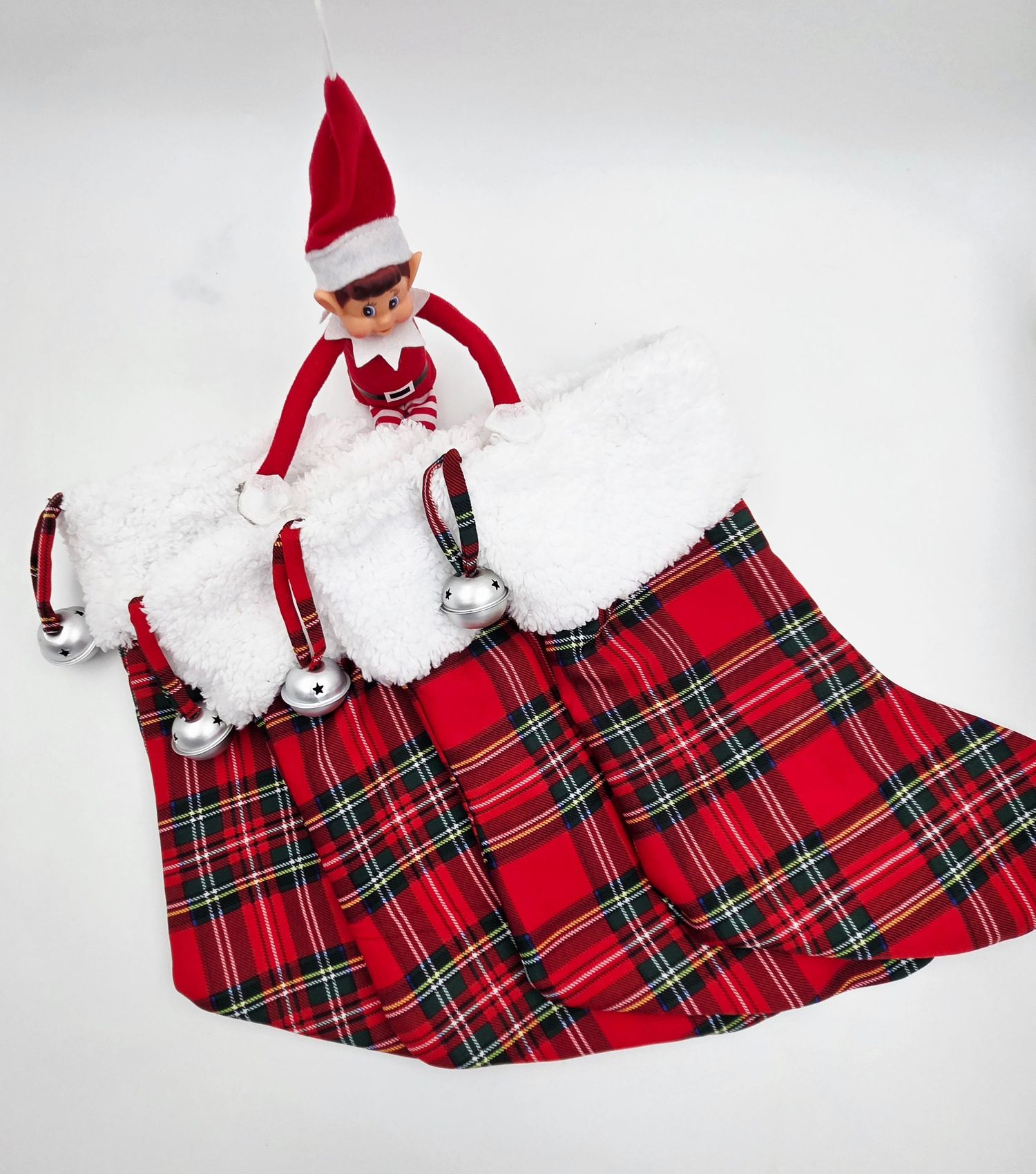 Elf on the Shelf doll sitting on a red plaid Christmas tree skirt with silver jingle bells and white fur trim.