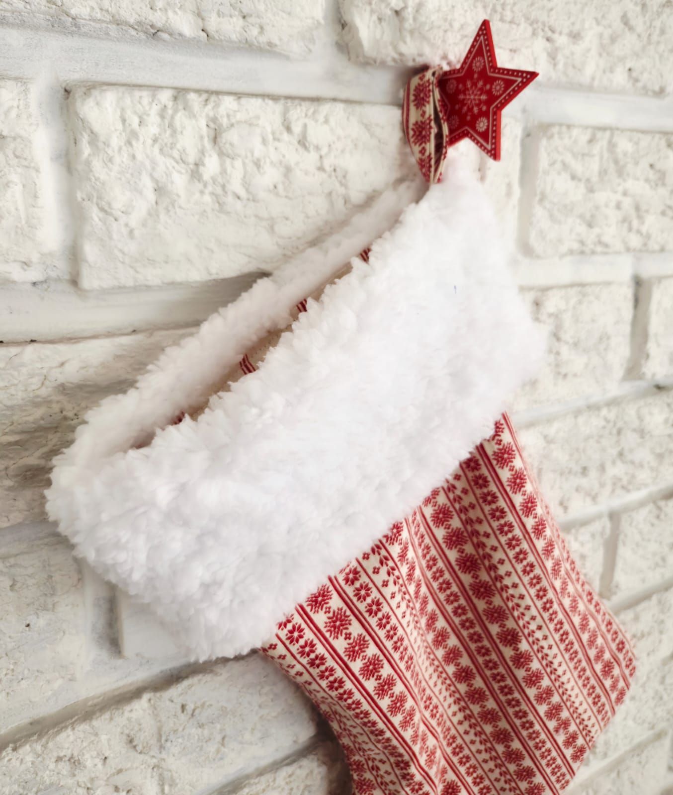 Red and white Nordic pattern Christmas stocking with faux fur cuff and red star hanger on white brick wall.