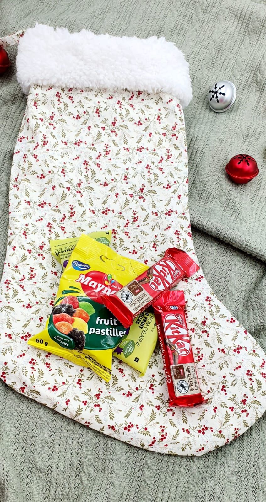 A festive Christmas stocking with holly print filled with KitKat bars and Maynards fruit pastilles candy beside jingle bells.