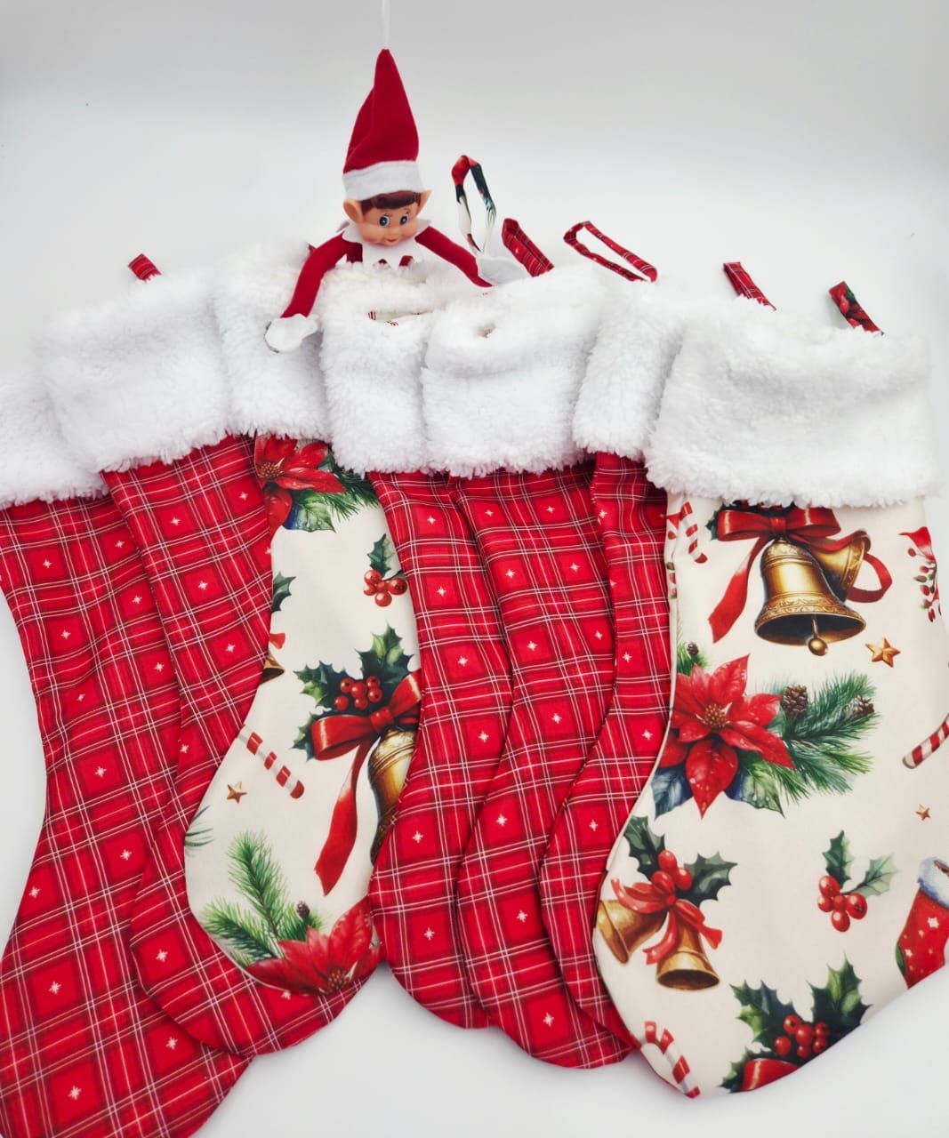 Four Christmas stockings with holiday prints and white trim displayed with an Elf on the Shelf peeking out from behind them.