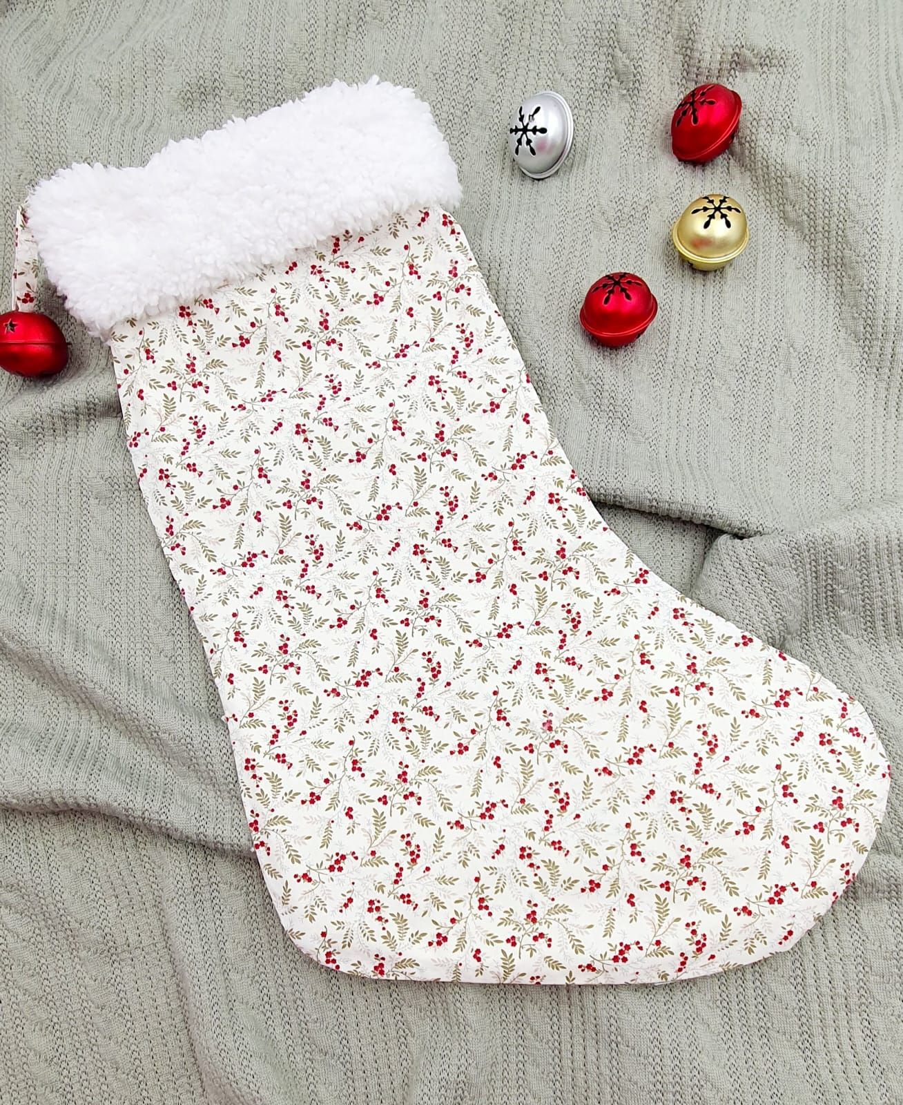 White Christmas stocking with red holly berry print pattern and fluffy white faux fur cuff, displayed on gray fabric.