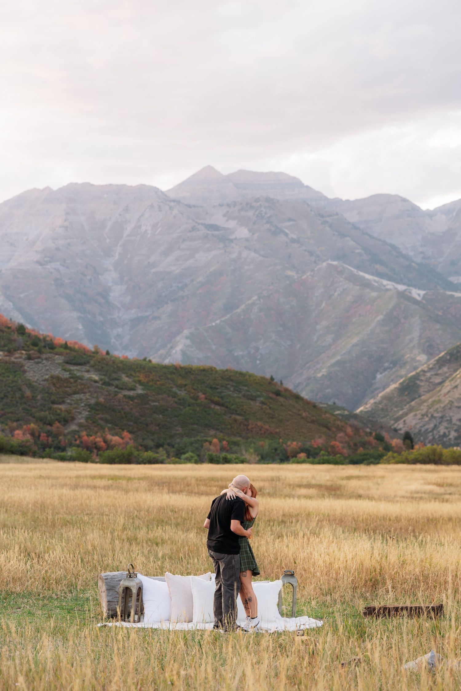 Couple embracing in golden field with stunning mountain backdrop during autumn outdoor photo session.