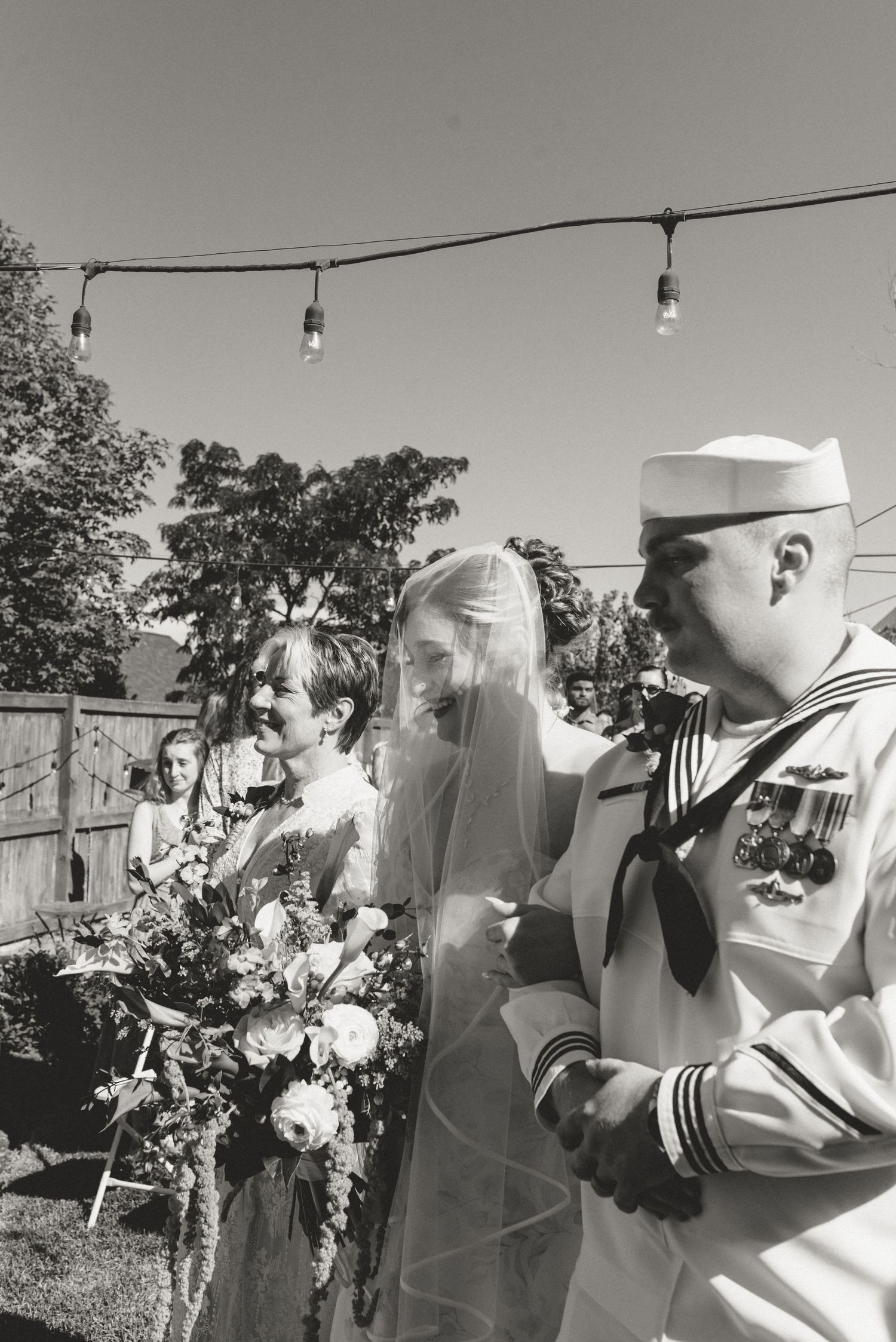 Black and white photo of a bride in veil with bouquet walking beside a Navy sailor in uniform at an outdoor wedding.
