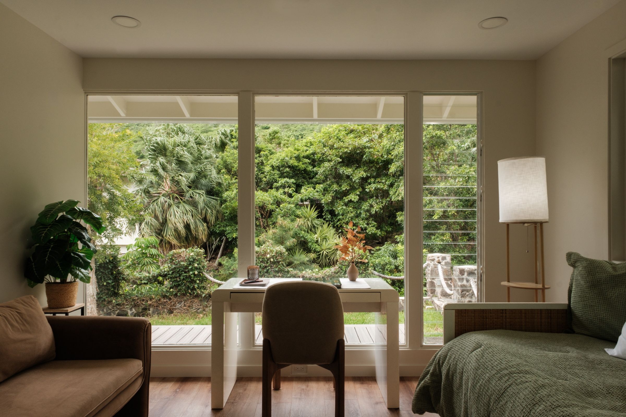 Home office with a desk and chair centered before garden-facing windows, a sofa, and a floor lamp.