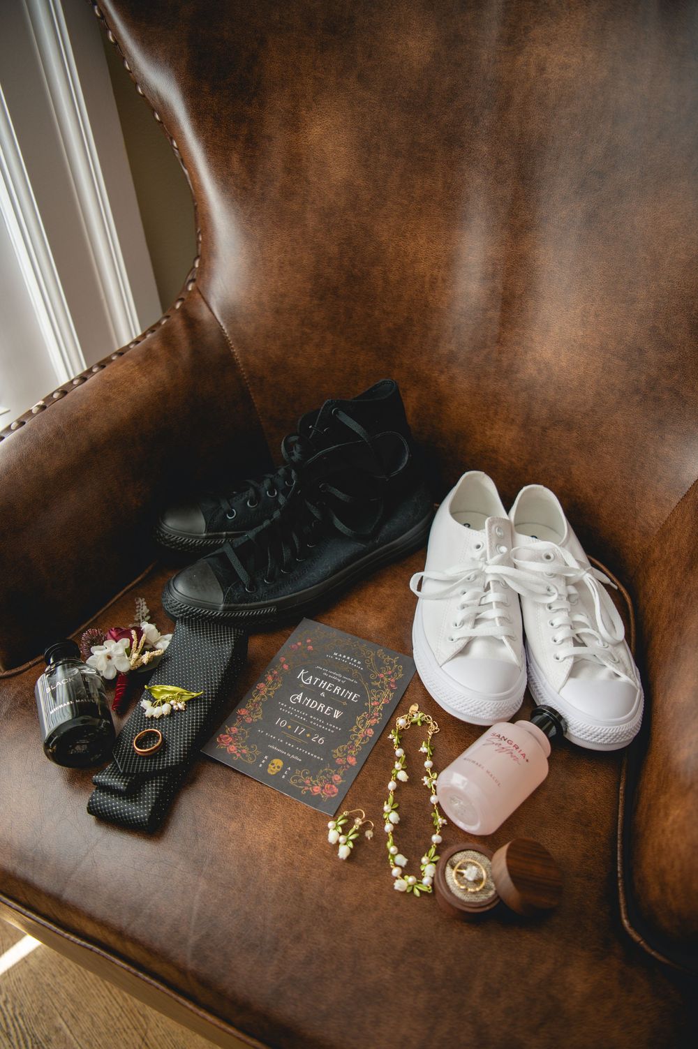 Black and white Converse sneakers, wedding invitation, boutonniere, perfume bottles and floral jewelry arranged on a brown leather chair.