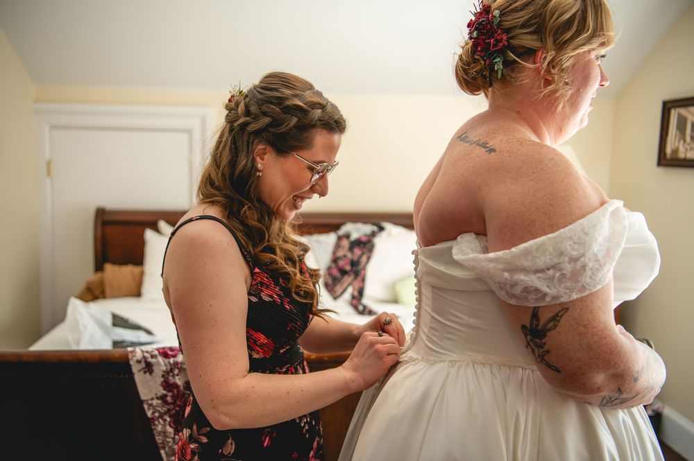 A woman in a floral dress helps a bride with a floral hair accessory fasten her off-the-shoulder white wedding gown in a bedroom.