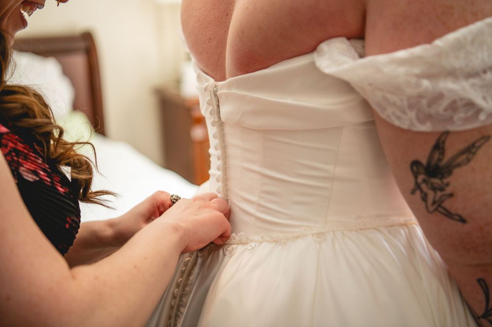 Bridesmaid in a floral dress laces up the back corset of a bride's white wedding gown, with a fairy tattoo visible on her shoulder.