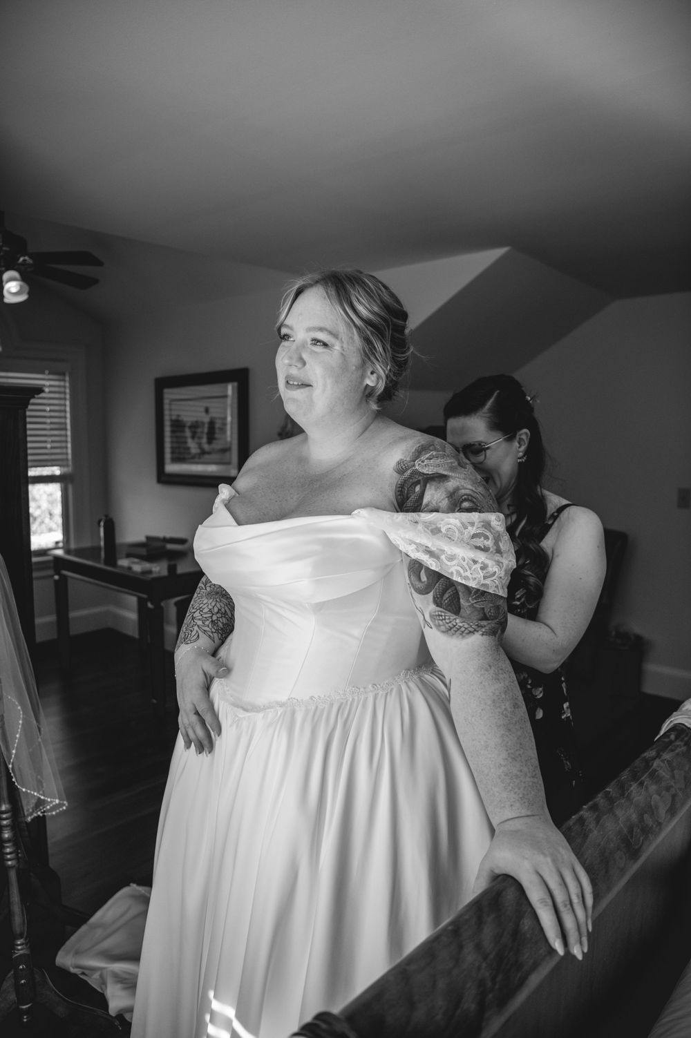 Black and white photo of a bride in an off-the-shoulder wedding gown getting ready as a woman helps fasten her dress in a bright attic room.