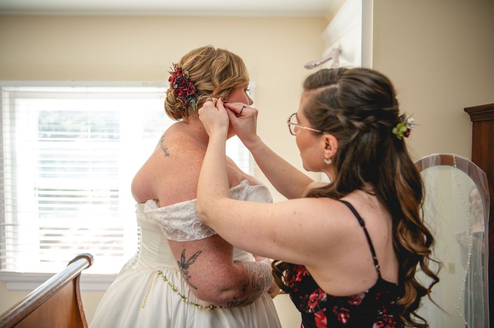 A bridesmaid in a floral dress helps a bride in an off-the-shoulder wedding gown fasten an earring before the ceremony.
