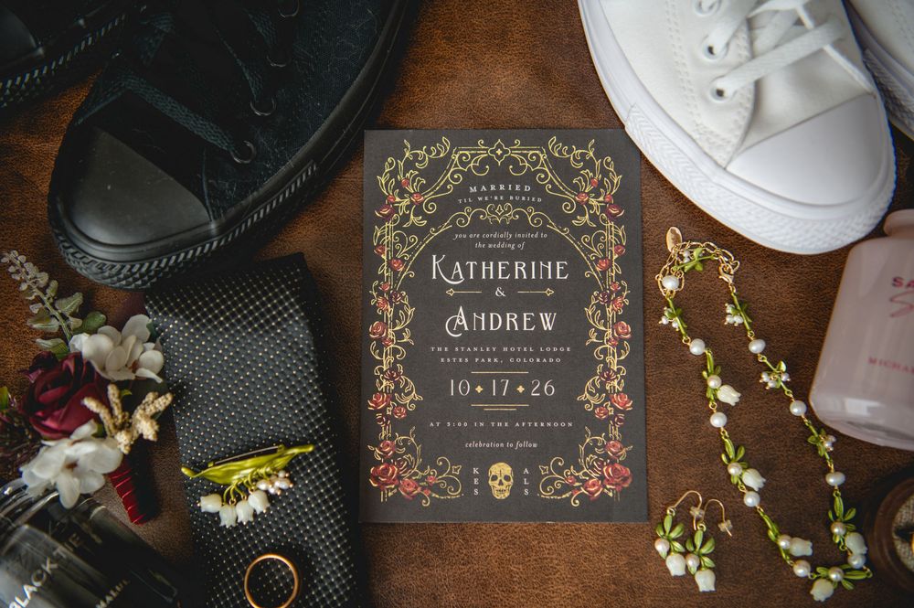 Dark floral wedding invitation for Katherine and Andrew on a wooden table with groom's shoes, necktie, boutonniere, white sneakers and pearl