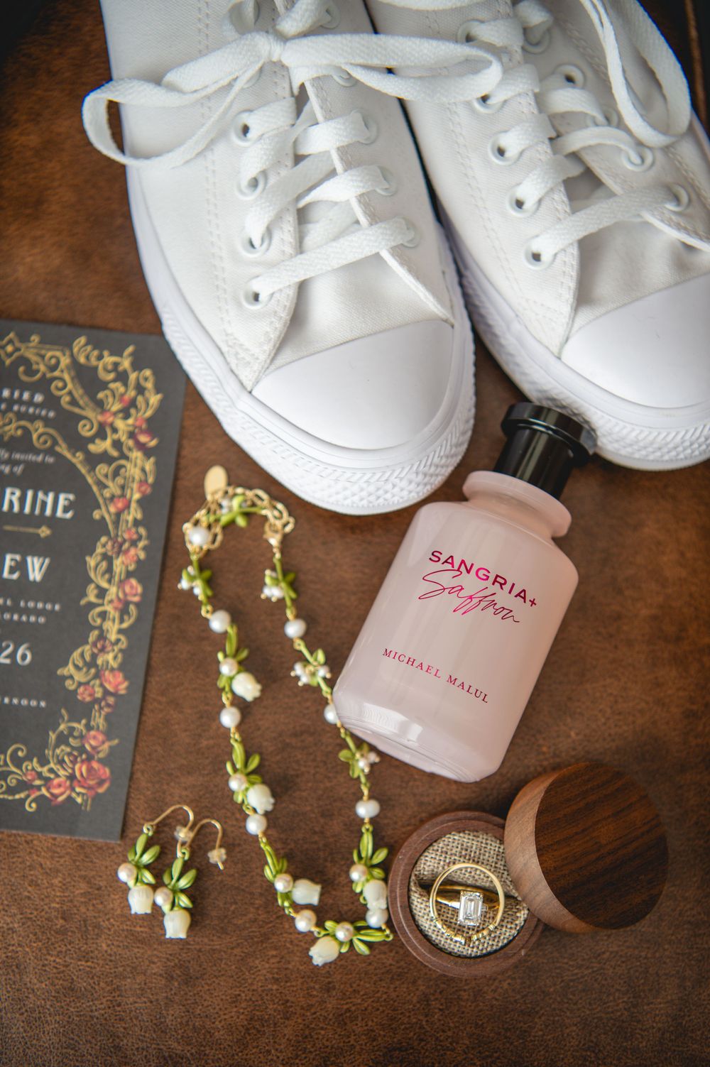 White canvas sneakers, floral bridal jewelry, a perfume bottle, wedding invitation and an engagement ring in a wooden box on a table.