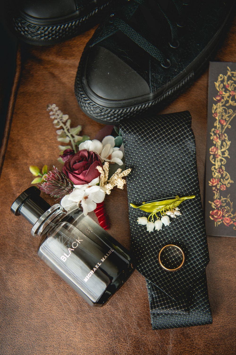 Wedding day flat lay with black sneakers, burgundy rose boutonniere, cologne bottle, black patterned tie and gold ring on brown leather.