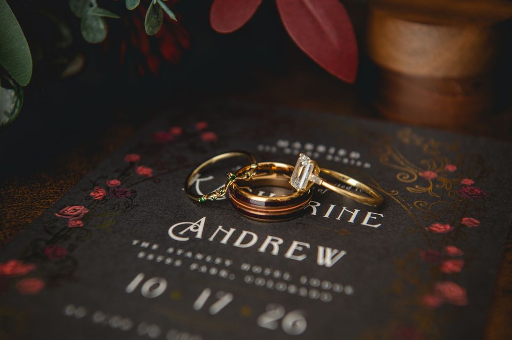 Gold wedding bands and a diamond engagement ring rest on a dark floral wedding invitation for Andrew, dated 10.17.26.