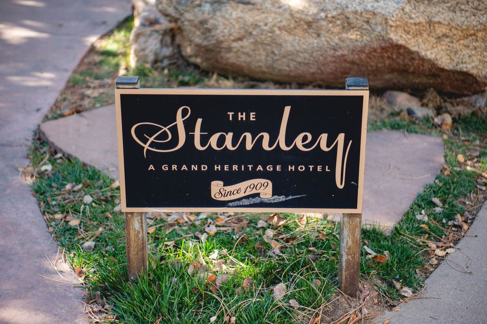 Wooden sign reading The Stanley, A Grand Heritage Hotel, Since 1909, set in a grassy area beside a large boulder and a paved walkway.