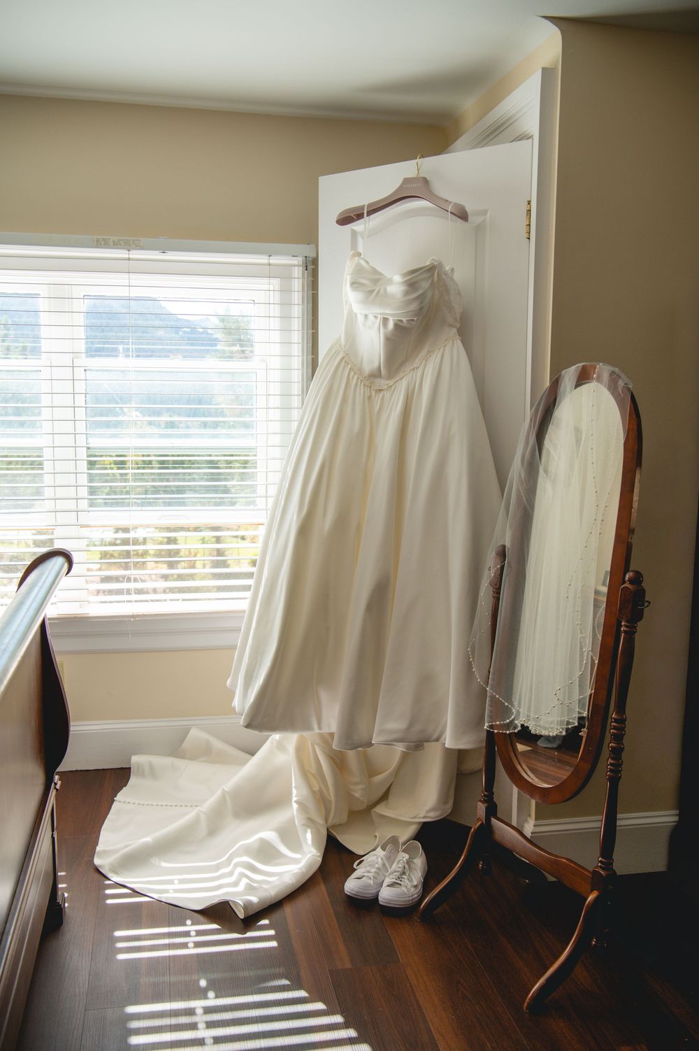 White strapless wedding dress with long train hanging on a door beside a veil on a mirror, with sneakers on the wood floor.