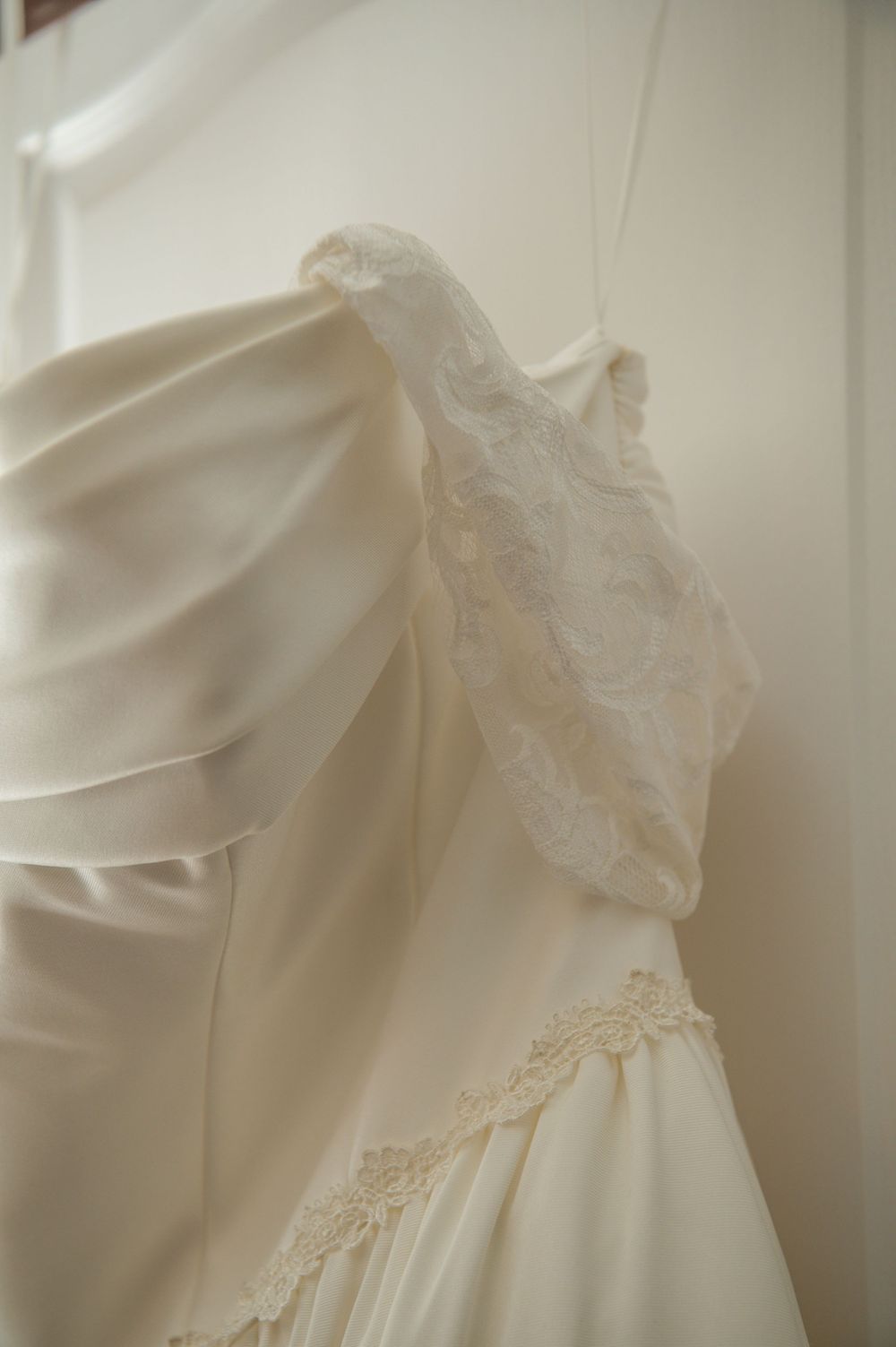 Ivory off-the-shoulder wedding dress with lace sleeve detail and satin bodice hanging against a soft white wall.