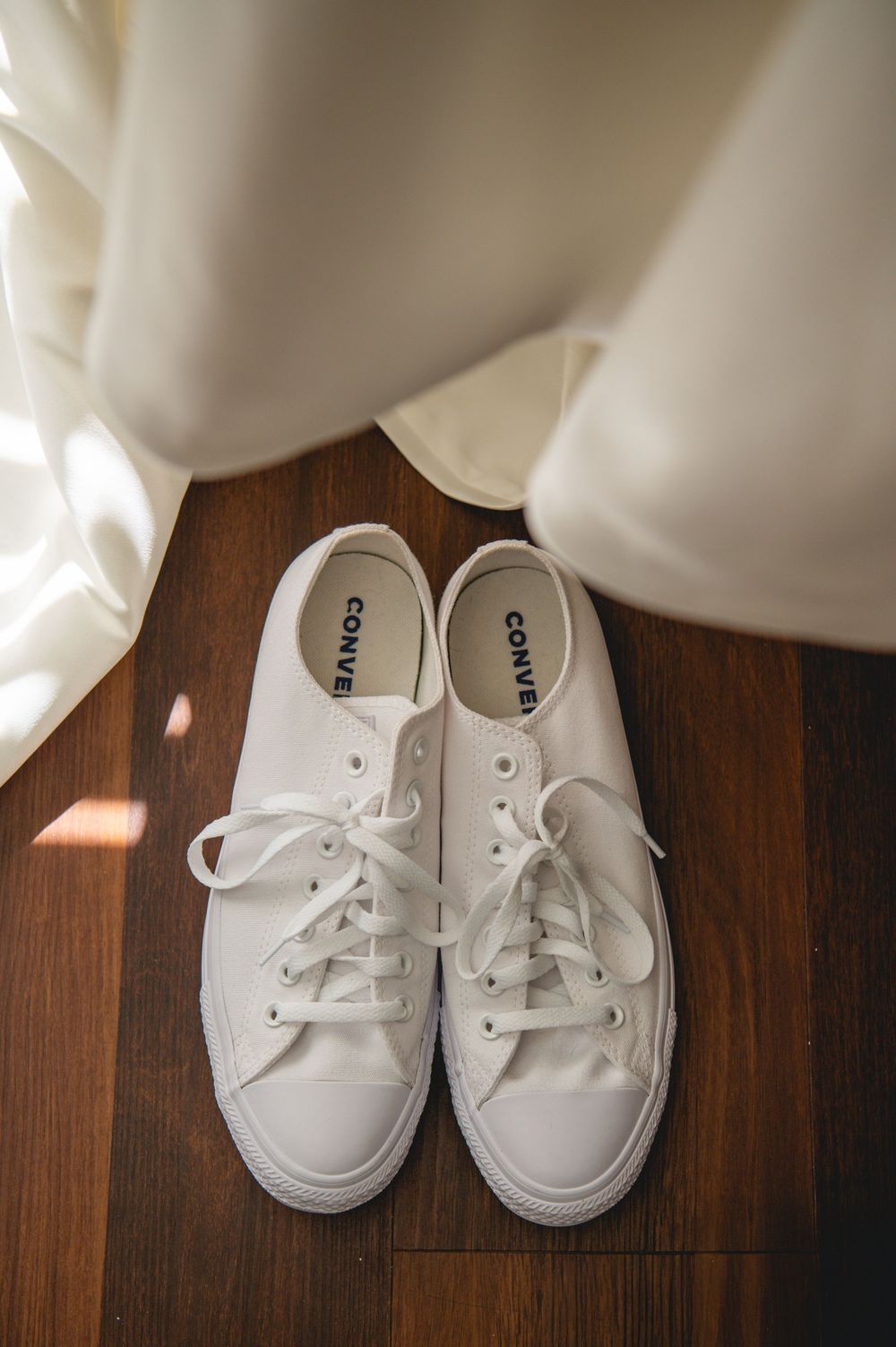 White Converse low-top sneakers with white laces on a wooden floor, partially covered by the hem of a flowing white wedding dress.