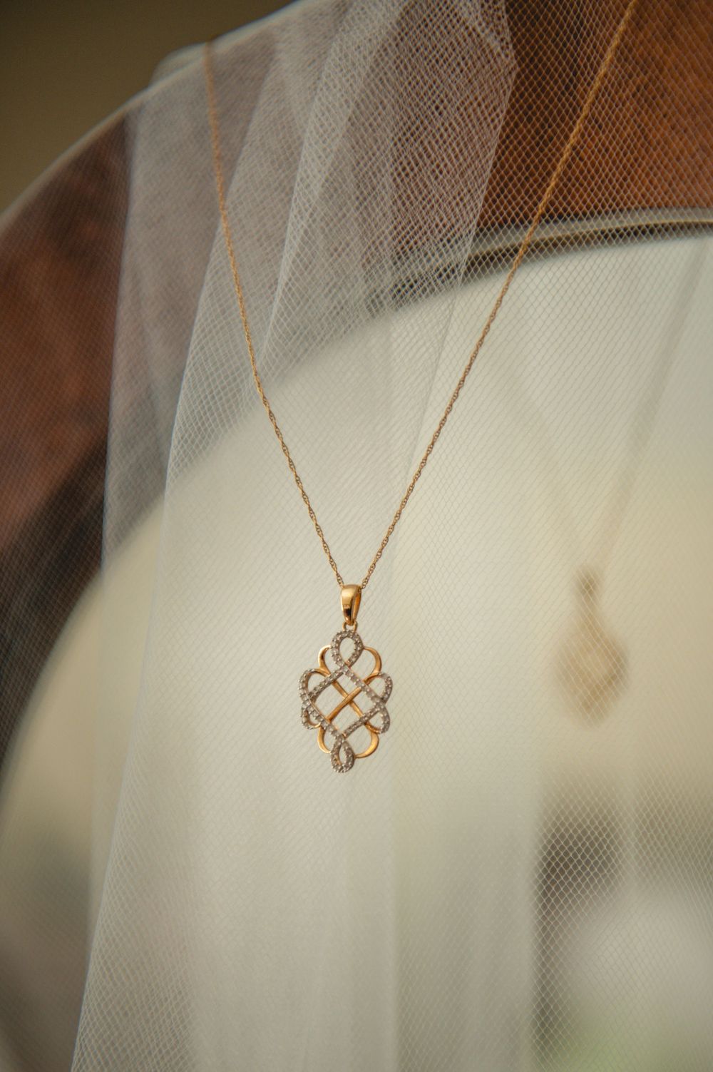 Gold pendant necklace with an intricate diamond-accented woven knot design hanging against a sheer white bridal veil.