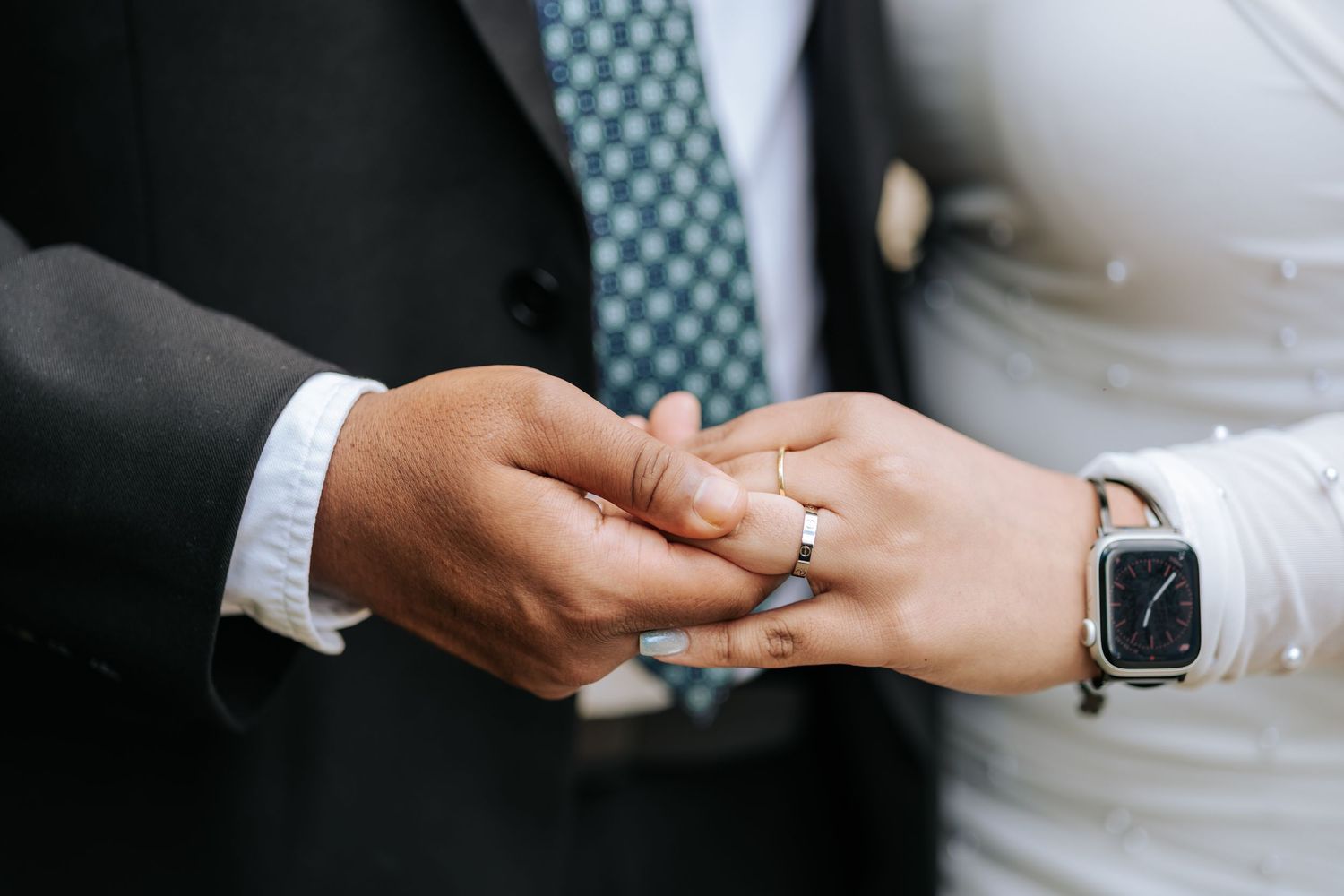 Close-up of a groom in a dark suit and green patterned tie holding hands with a bride wearing a smartwatch during a wedding ring exchange.