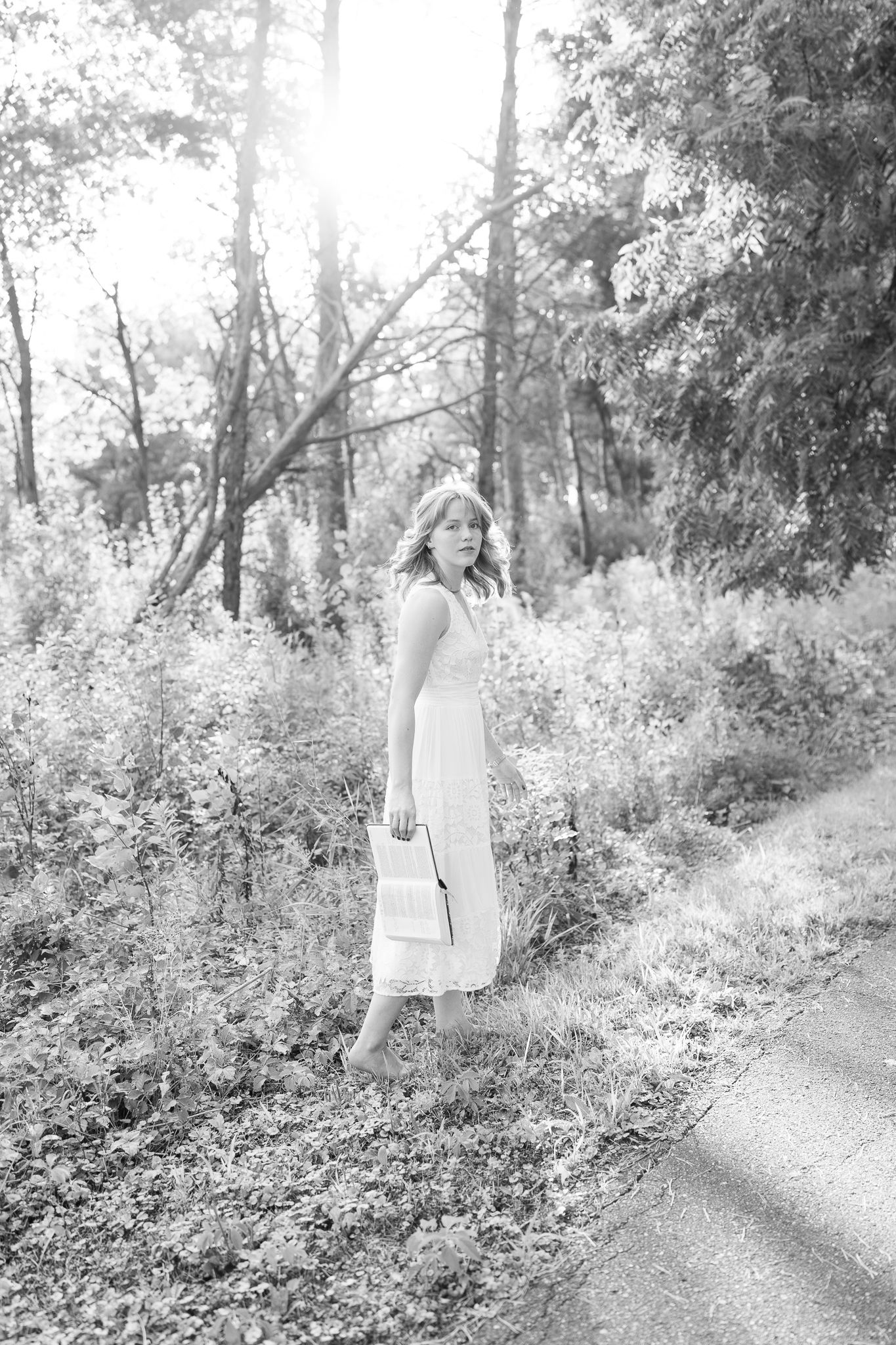A black and white photo of a woman in a white dress holding a purse on a sunlit wooded path.