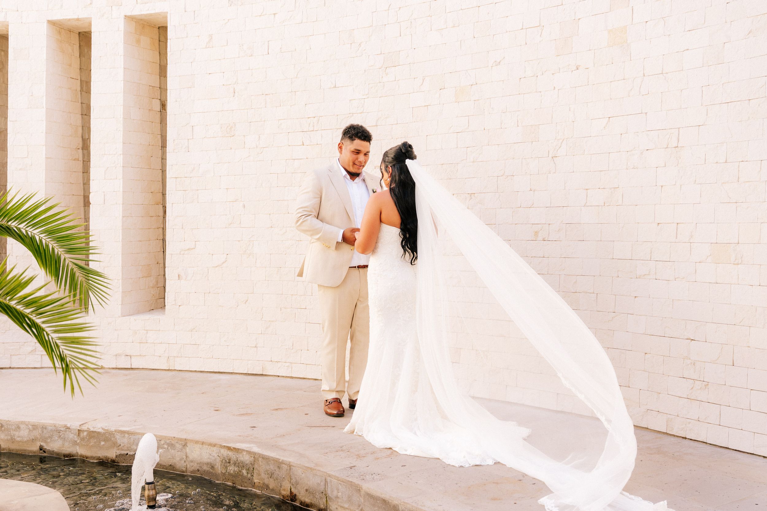 Bride in a white gown with a long flowing veil embraces her groom in a cream suit beside a fountain against a white stone wall.