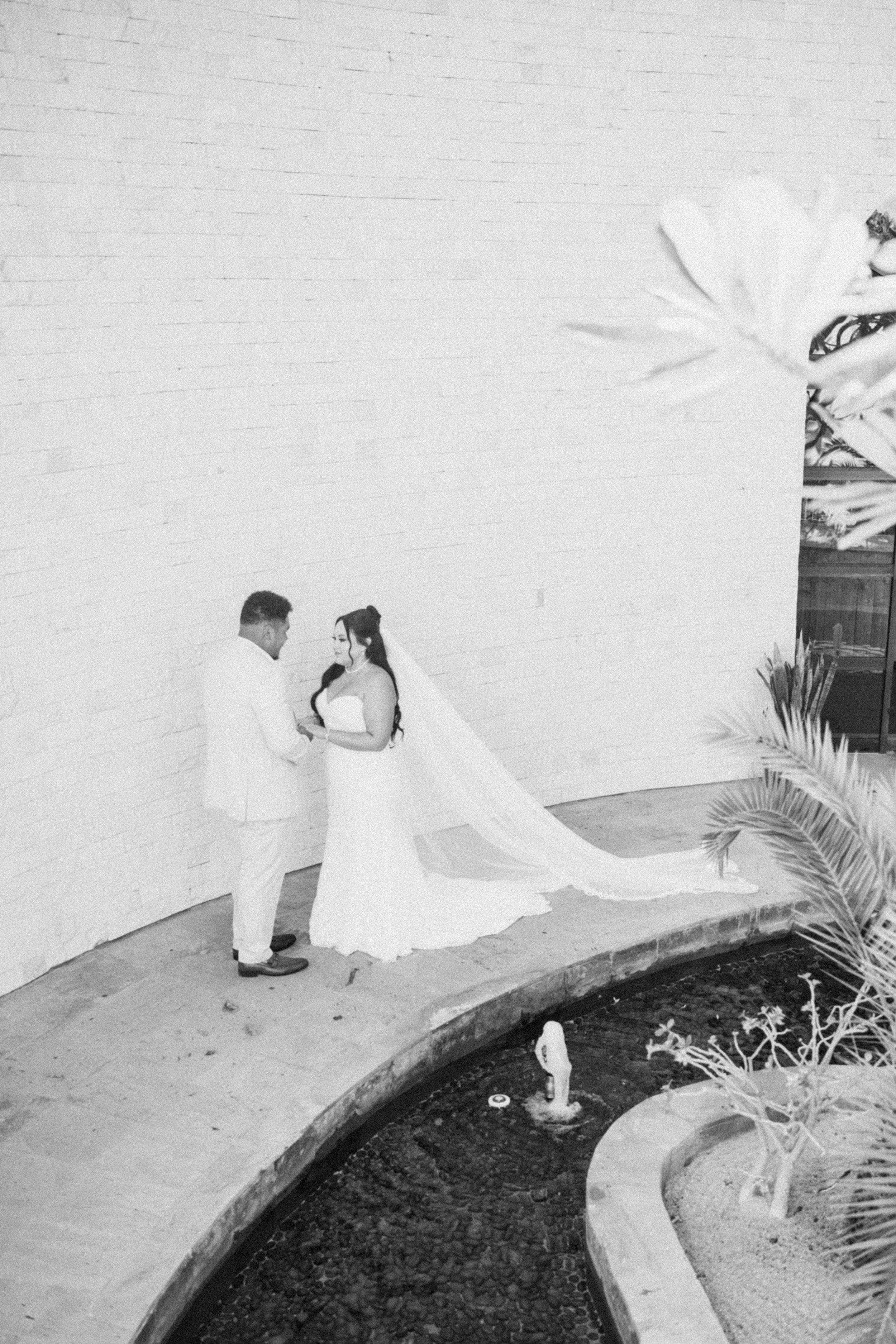 Black and white photo of a bride in a long veil and a groom in a light suit holding hands beside a curved pond and white brick wall.