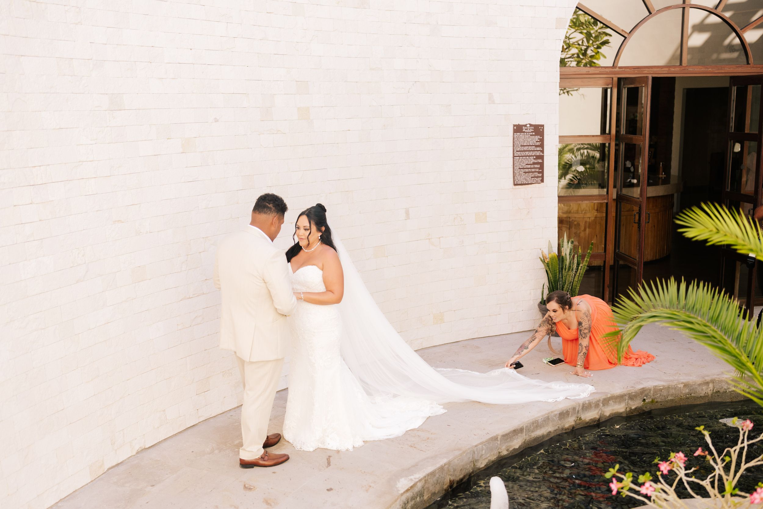 A bride in a white gown with a long veil stands beside a groom in a cream suit as a bridesmaid in orange arranges her train near a tropical 